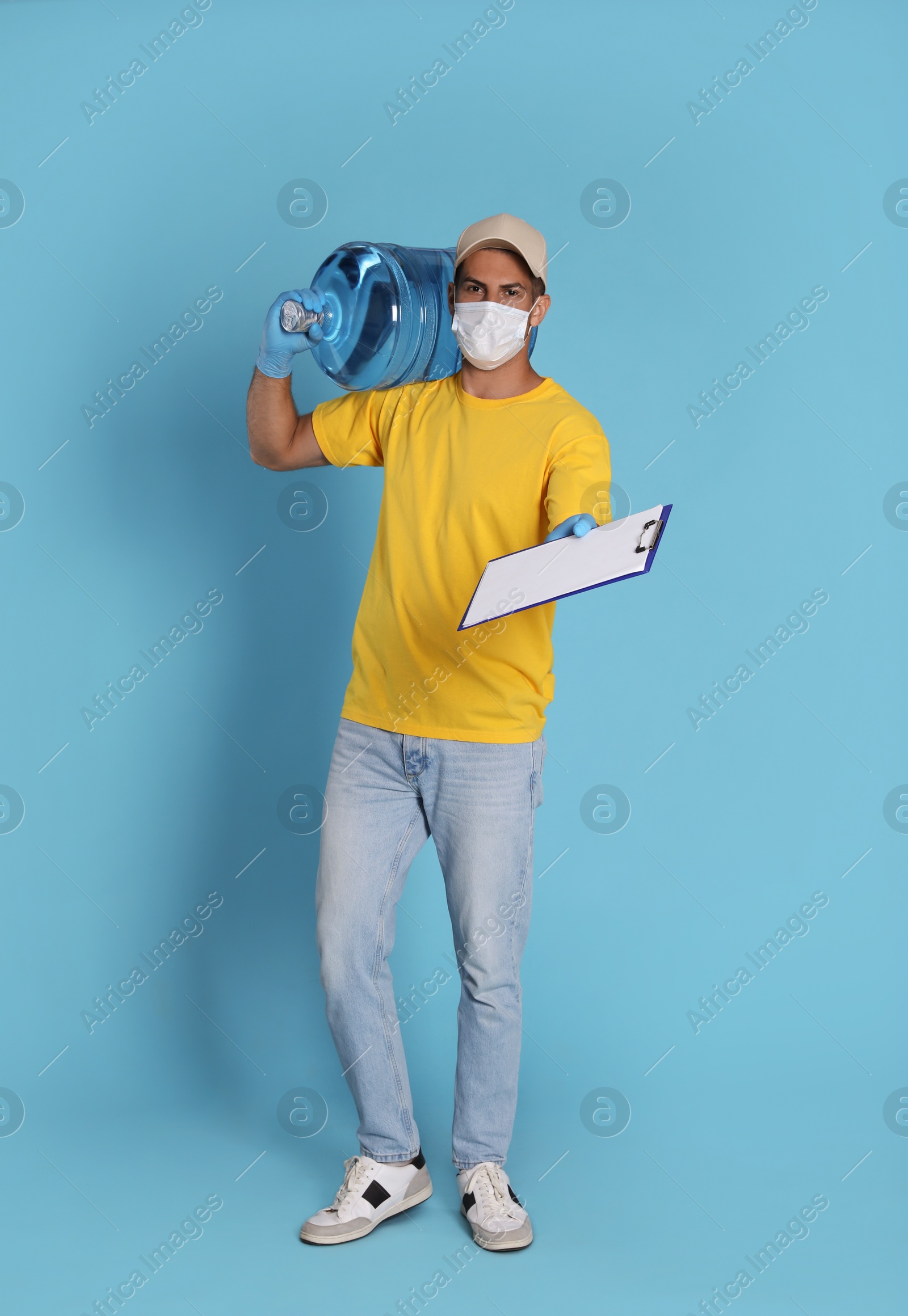 Photo of Courier in medical mask with bottle for water cooler and clipboard on light blue background. Delivery during coronavirus quarantine