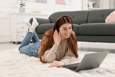 Photo of Happy woman with laptop and headphones having video chat on rug in living room