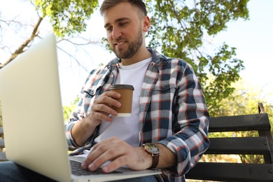 Young man with paper cup of coffee working on laptop in park