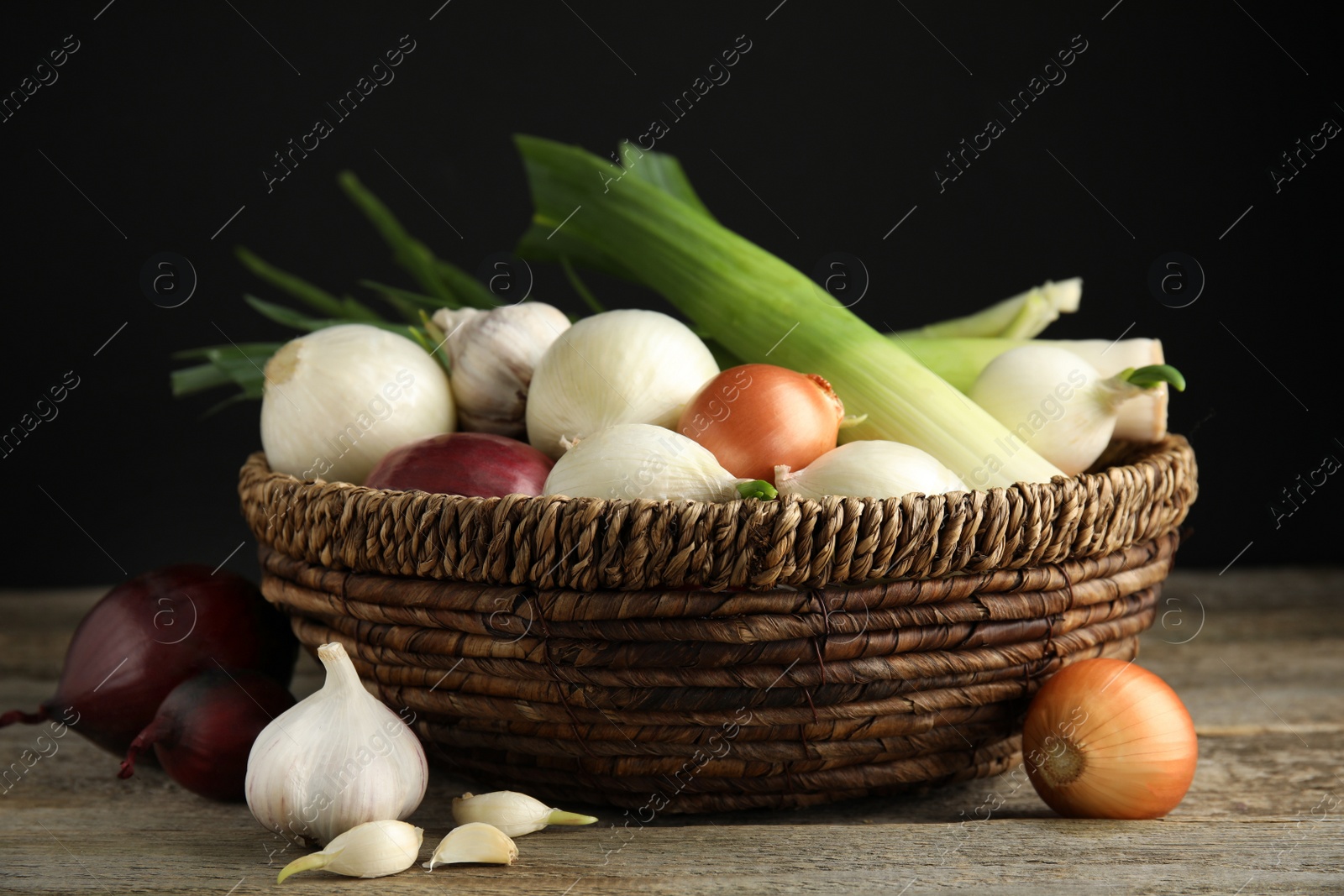 Photo of Wicker basket with fresh onion bulbs, leeks and garlic on wooden table