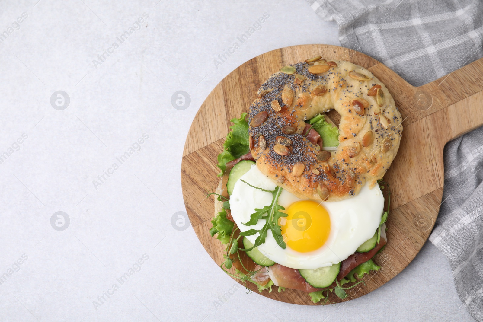 Photo of Tasty bagel with cured ham, egg, cucumber and salad mix on light table, top view. Space for text