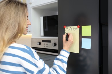 Photo of Young woman writing To do list on refrigerator door in kitchen