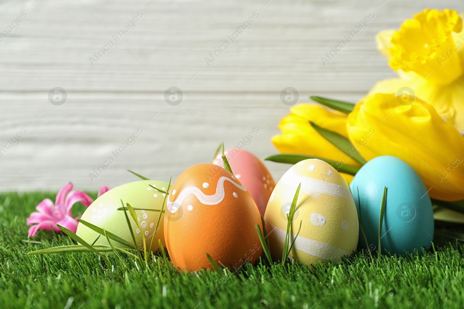 Photo of Colorful painted Easter eggs and flowers on green grass against wooden background, space for text