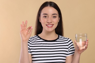 Photo of Happy woman with glass of water showing pill on beige background