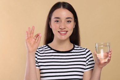 Happy woman with glass of water showing pill on beige background