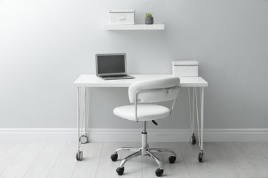 Photo of Stylish workplace with laptop and comfortable chair near white wall indoors. Interior design