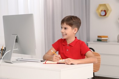 Boy writing in notepad while using computer at desk in room. Home workplace