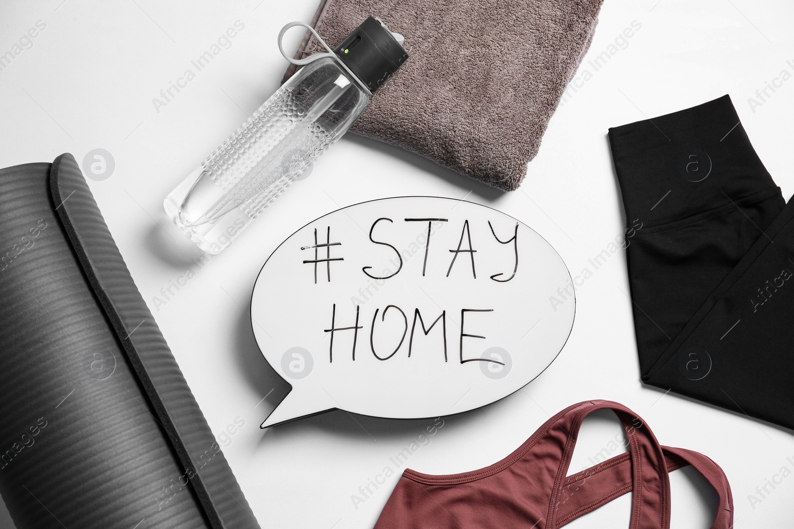 Photo of Stylish sportswear, yoga mat and speech bubble with hashtag Stay at Home on white background, flat lay. Self isolation during COVID‑19 pandemic