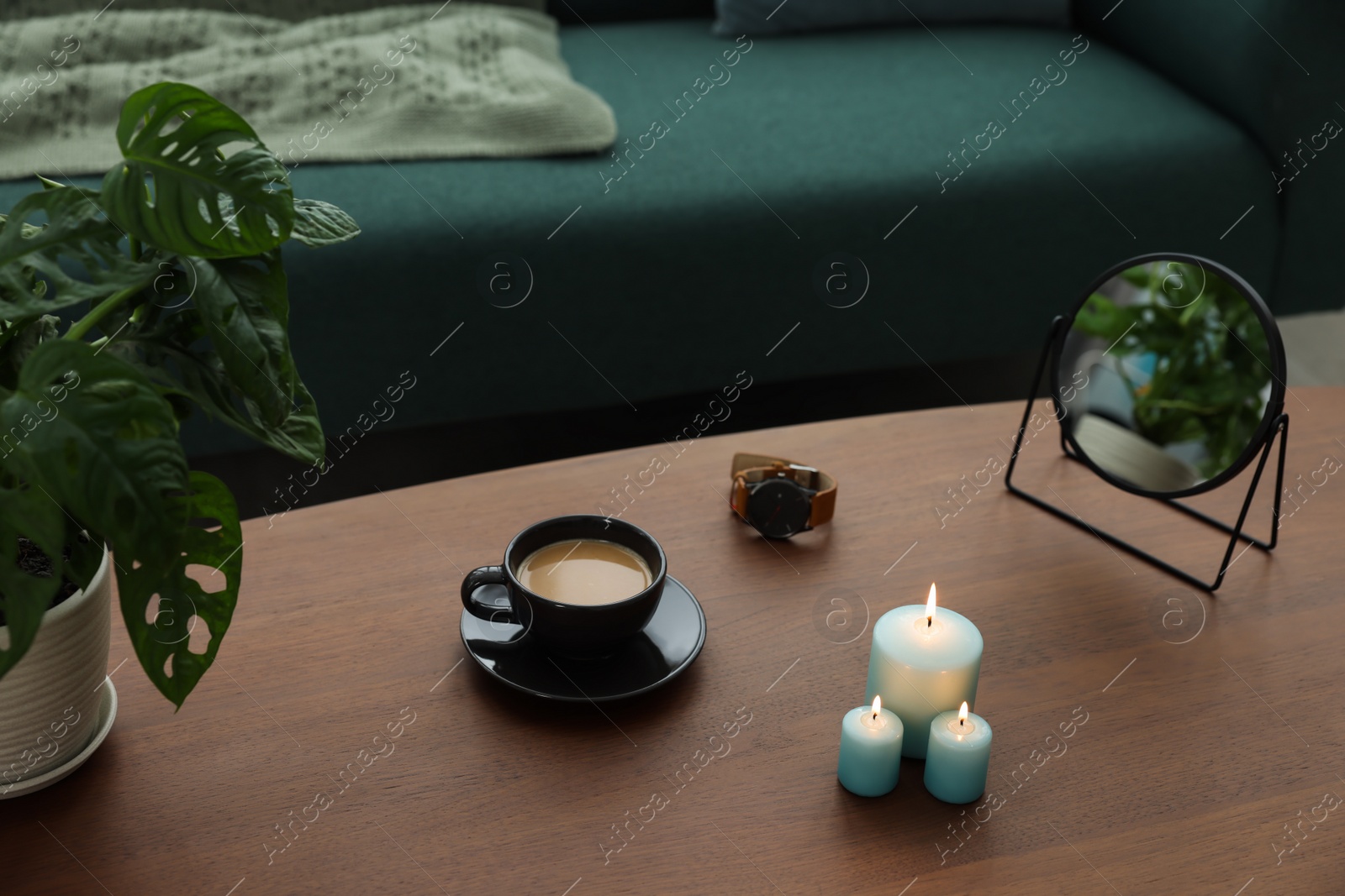 Photo of Wooden table with cup of coffee, candles, mirror, wristwatch and houseplant near sofa in room. Interior design