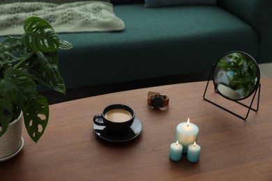 Wooden table with cup of coffee, candles, mirror, wristwatch and houseplant near sofa in room. Interior design