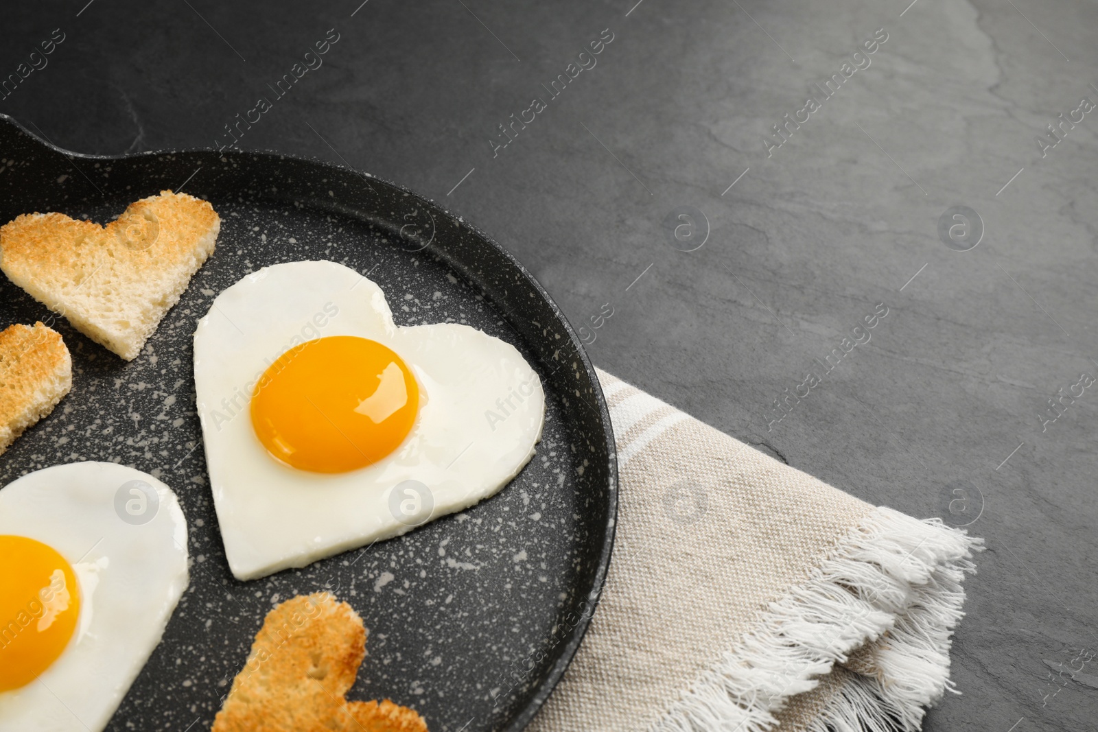 Photo of Heart shaped fried eggs and toasts in frying pan on grey table, closeup. Space for text