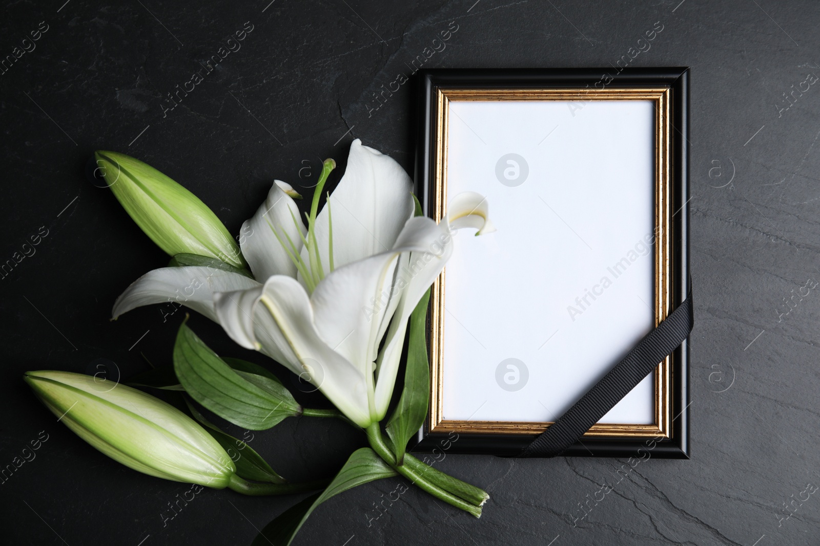 Photo of Funeral photo frame with ribbon and white lily on black table, flat lay. Space for design