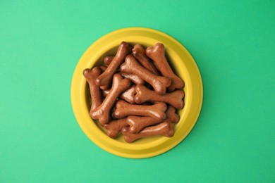 Photo of Yellow bowl with bone shaped dog cookies on green background, top view