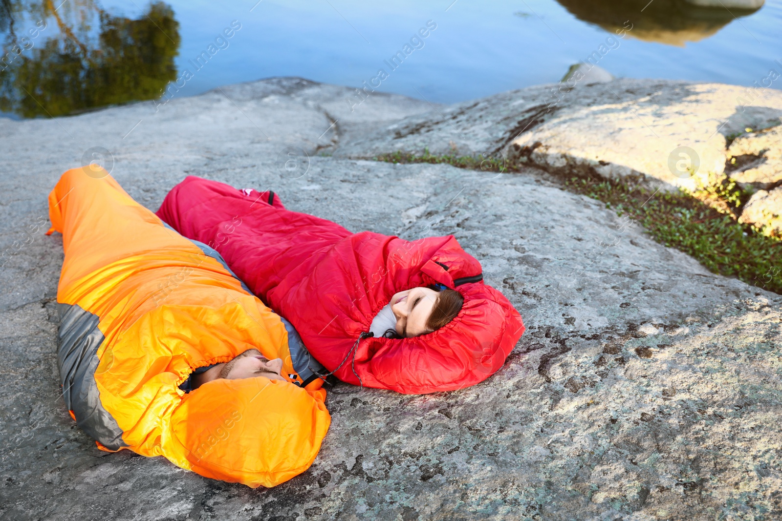 Photo of Young couple resting in sleeping bags on cliff near lake