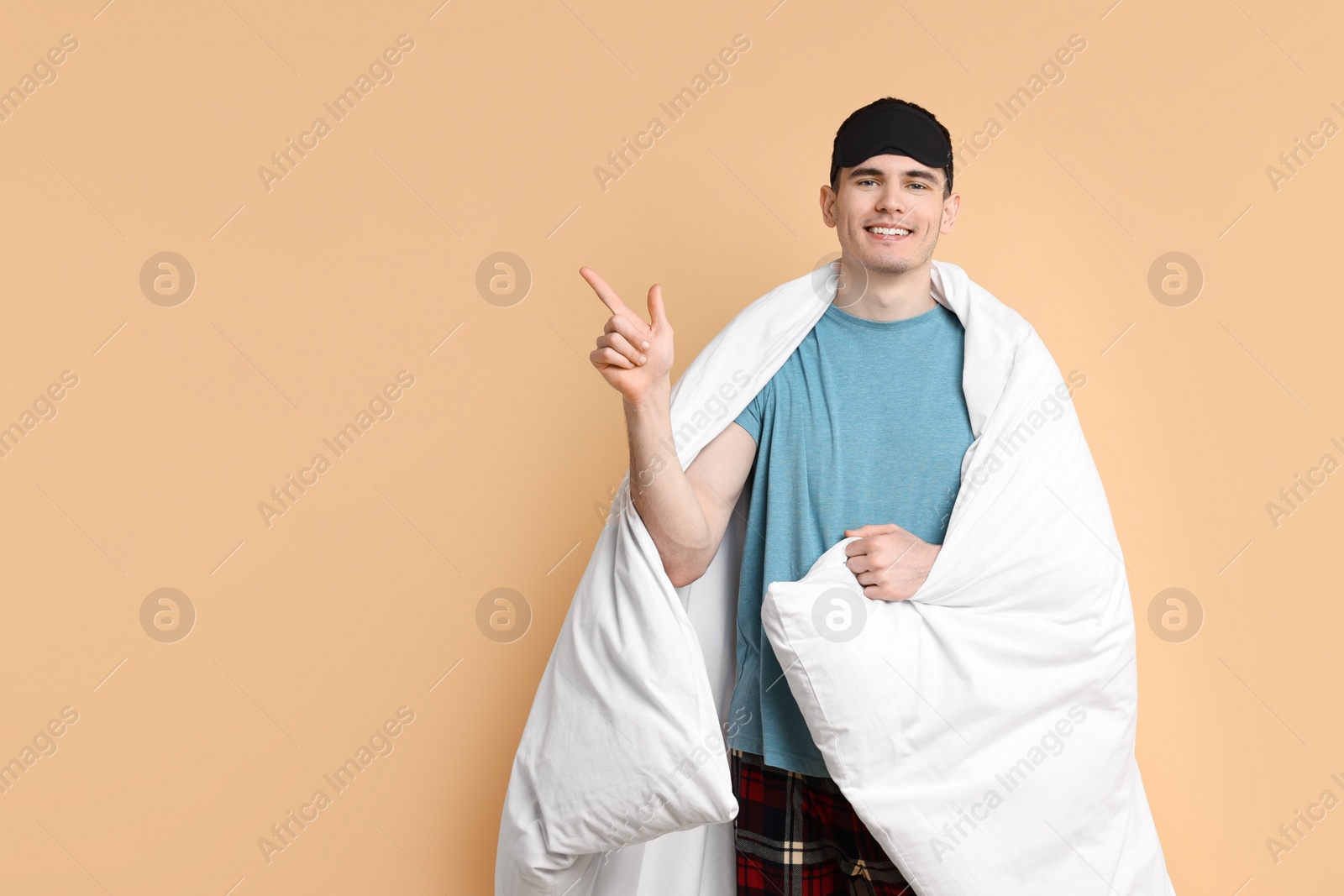 Photo of Happy man in pyjama and sleep mask wrapped in blanket on beige background, space for text