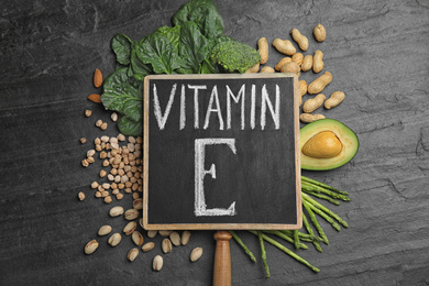 Photo of Small chalkboard with phrase Vitamin E and different products on dark background, flat lay