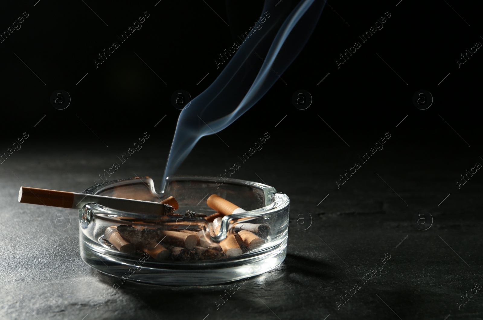 Photo of Smoldering cigarette in glass ashtray on grey table against black background. Space for text