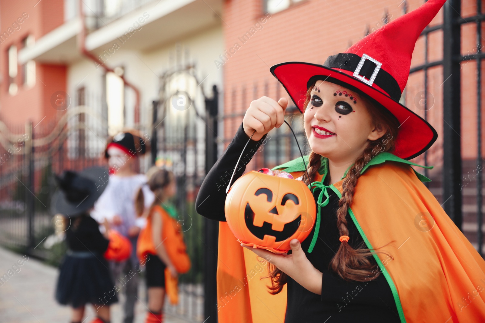 Photo of Cute little girl with pumpkin candy bucket wearing Halloween costume outdoors