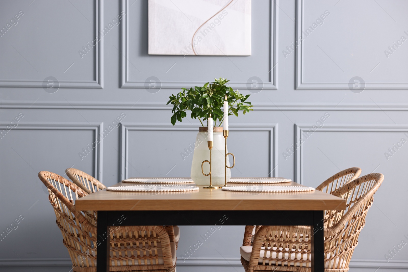 Photo of Rattan chairs, table, burning candles and vase with green branches in stylish dining room