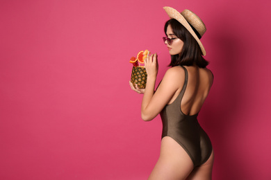 Beautiful young woman with exotic cocktail wearing swimsuit, hat and sunglasses on pink background. Space for text
