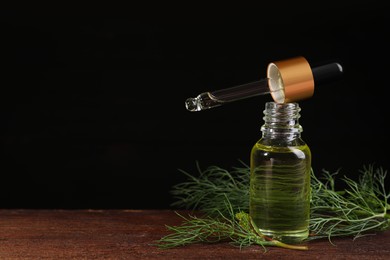 Photo of Bottle of essential oil, pipette and fresh dill on wooden table against black background, closeup. Space for text