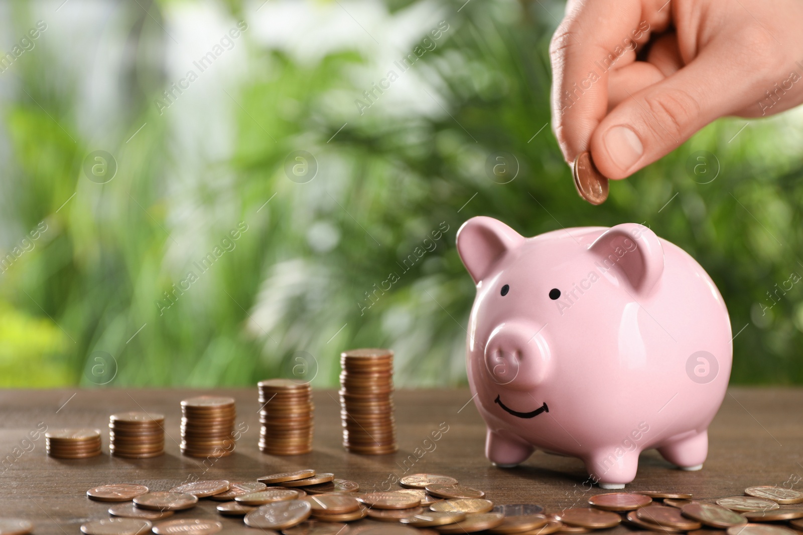 Photo of Woman putting coin into piggy bank at wooden table against blurred green background, closeup. Space for text