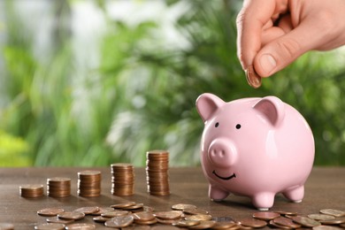 Photo of Woman putting coin into piggy bank at wooden table against blurred green background, closeup. Space for text