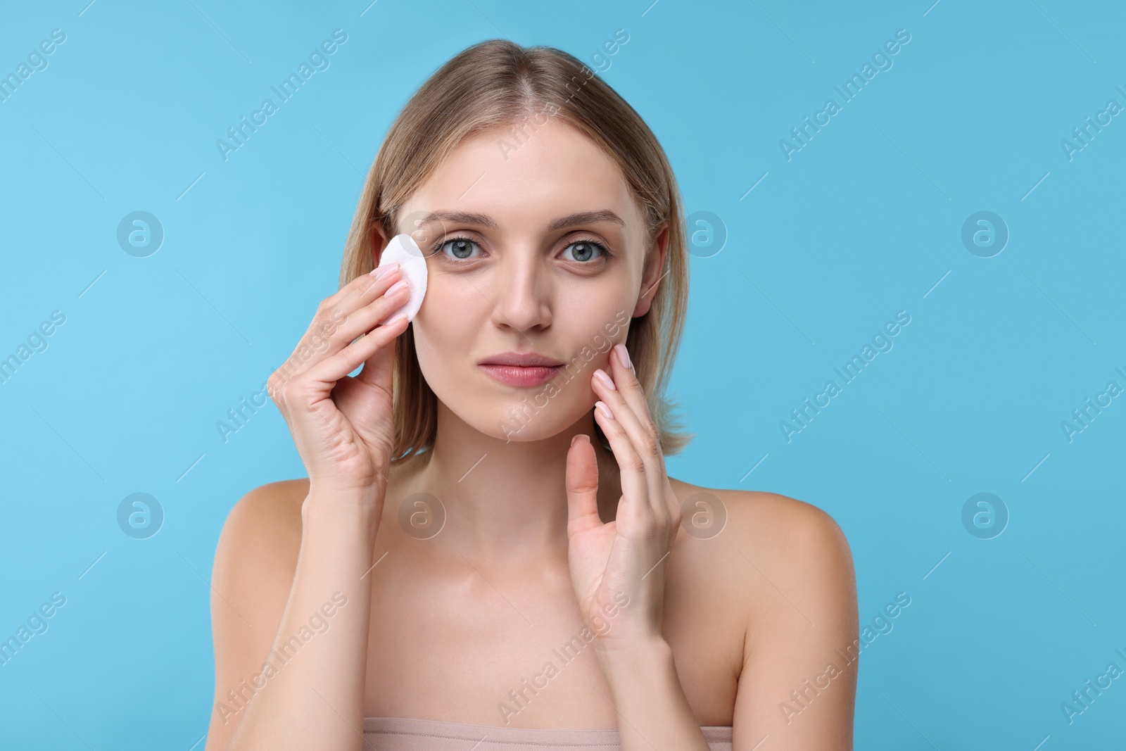 Photo of Young woman cleaning face with cotton pad on light blue background