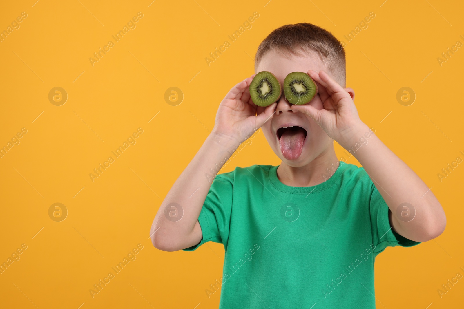 Photo of Emotional boy covering eyes with halves of fresh kiwi and showing tongue on orange background, space for text