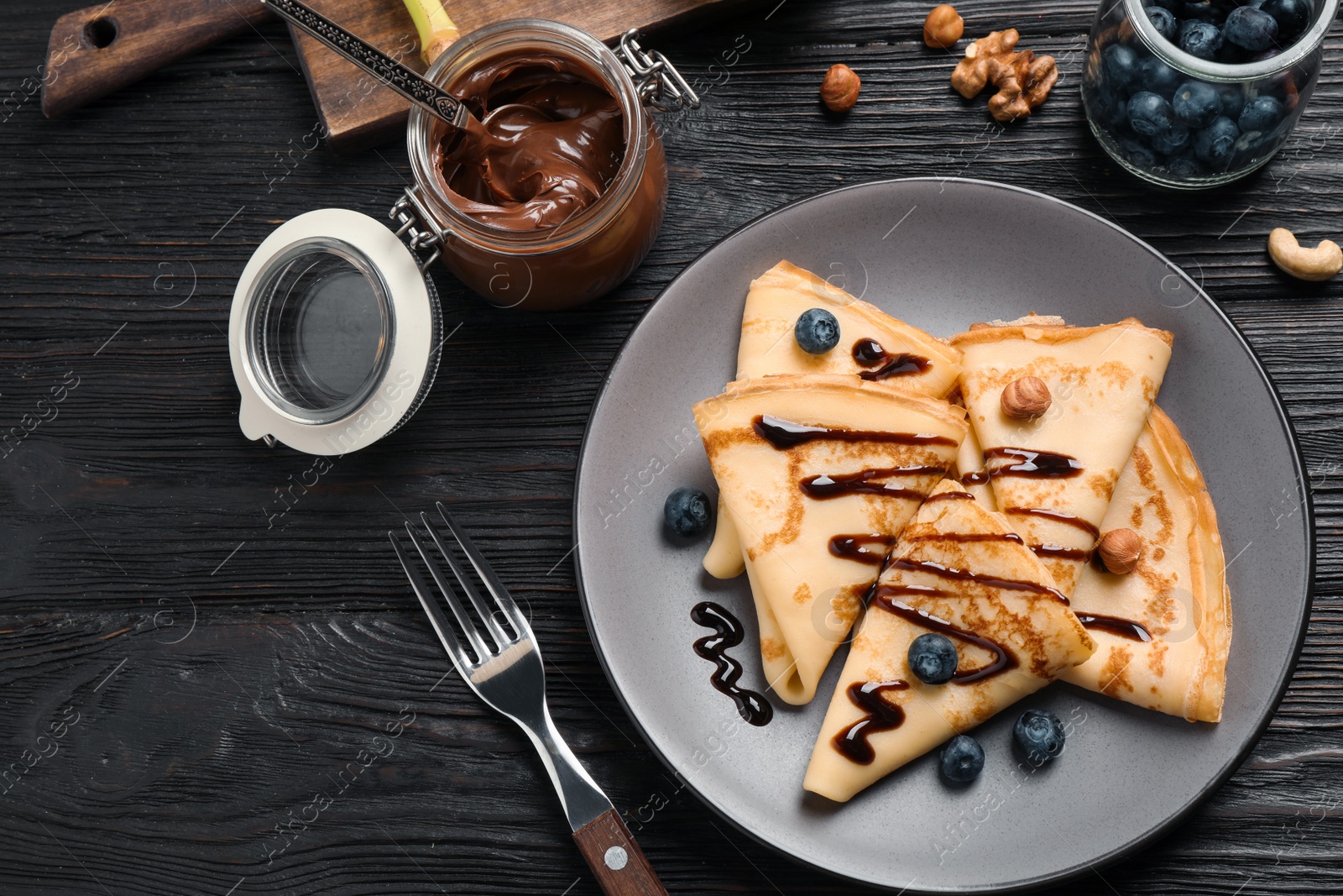 Photo of Delicious thin pancakes with chocolate, blueberries and nuts on black wooden table, flat lay