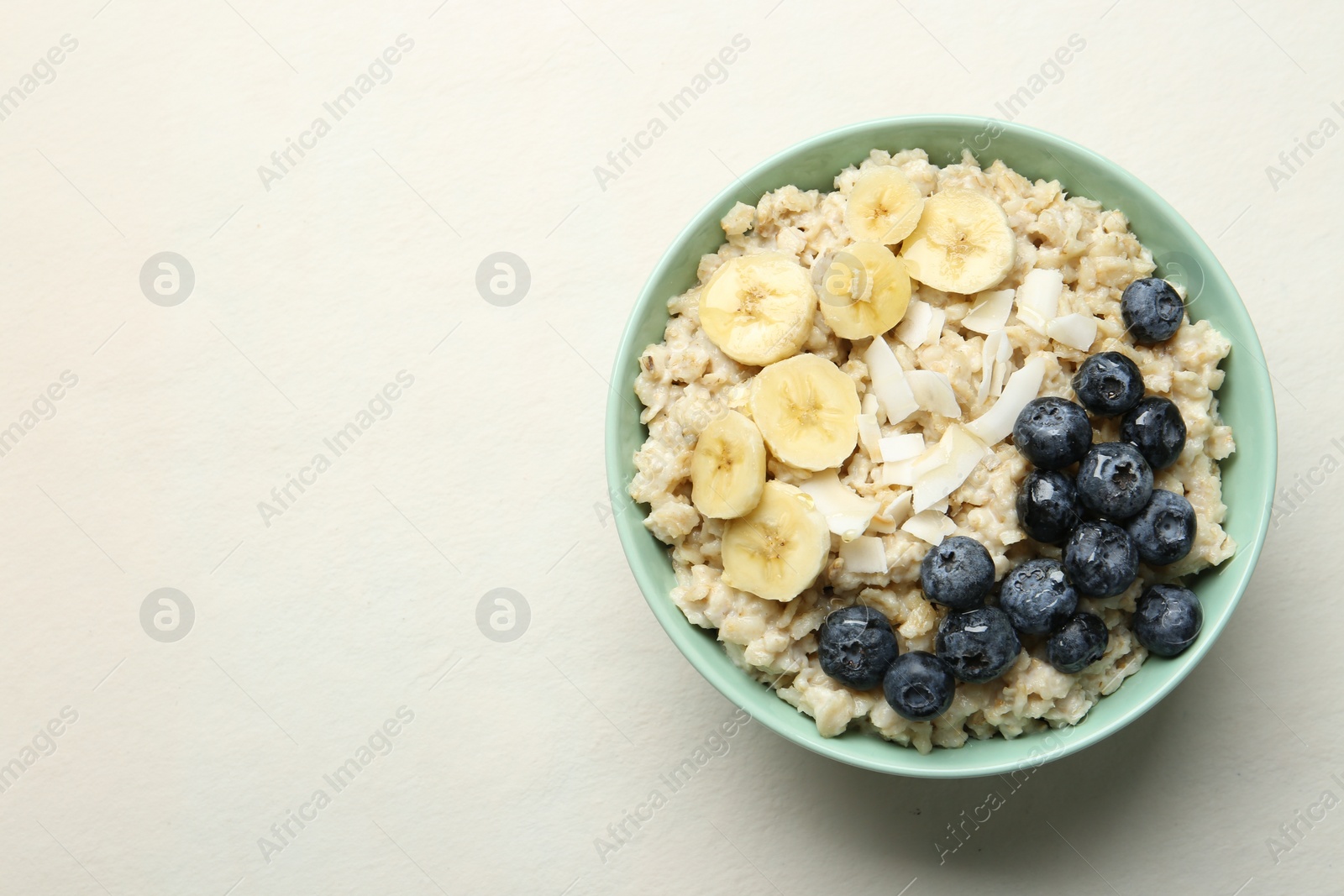 Photo of Tasty oatmeal with banana, blueberries, coconut flakes and honey served in bowl on beige table, top view. Space for text