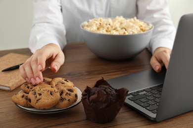 Photo of Bad habits. Woman eating cookies while using laptop at wooden table, closeup