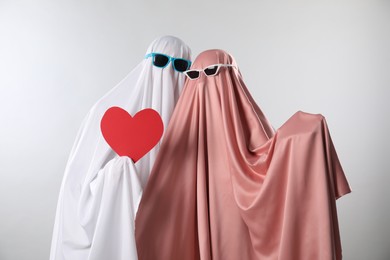 Photo of Cute ghosts. Couple in color sheets and sunglasses with red heart on light grey background
