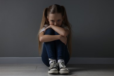 Sad girl sitting on floor near dark grey wall indoors