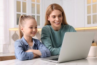 Happy woman and her daughter with laptop at white table indoors