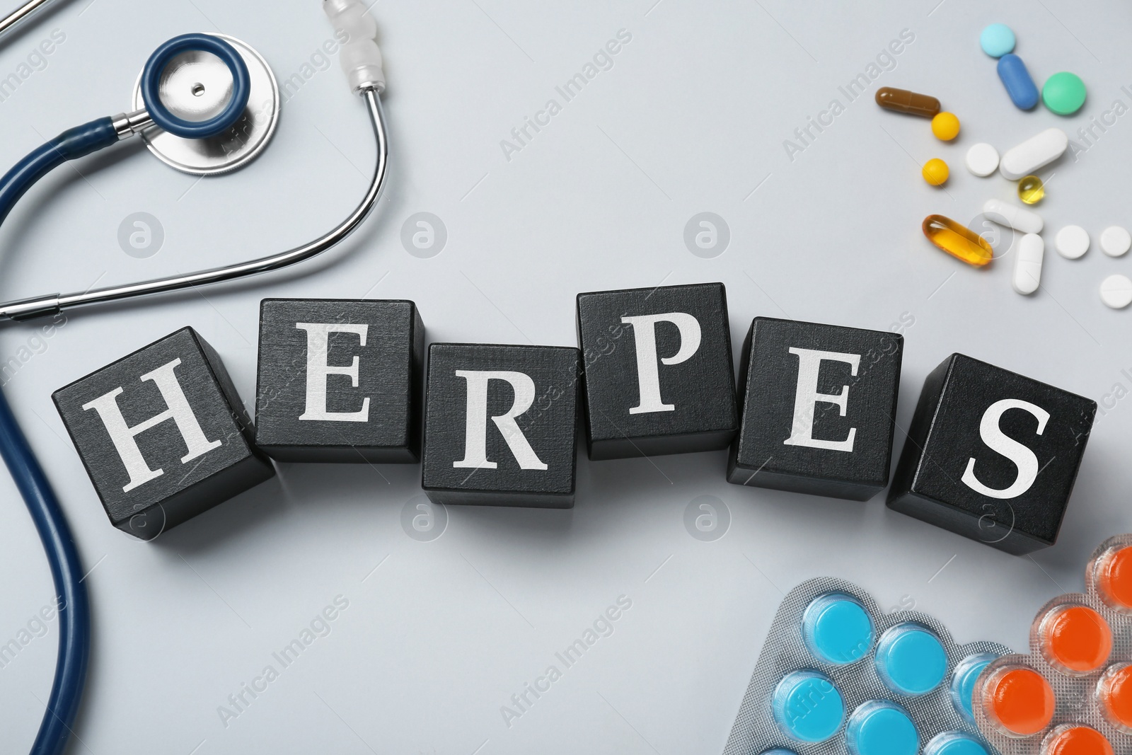 Photo of Word Herpes made of black wooden cubes, different pills and stethoscope on grey background, flat lay