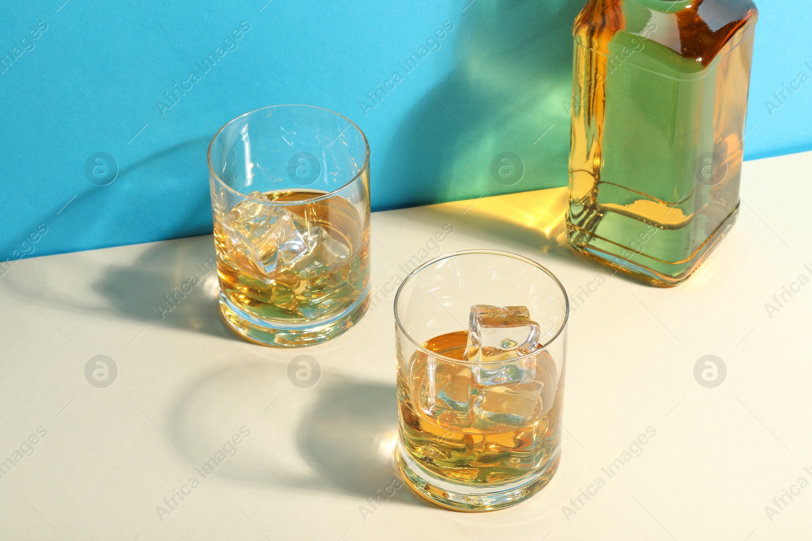 Photo of Whiskey with ice cubes in glasses and bottle on white table against light blue background