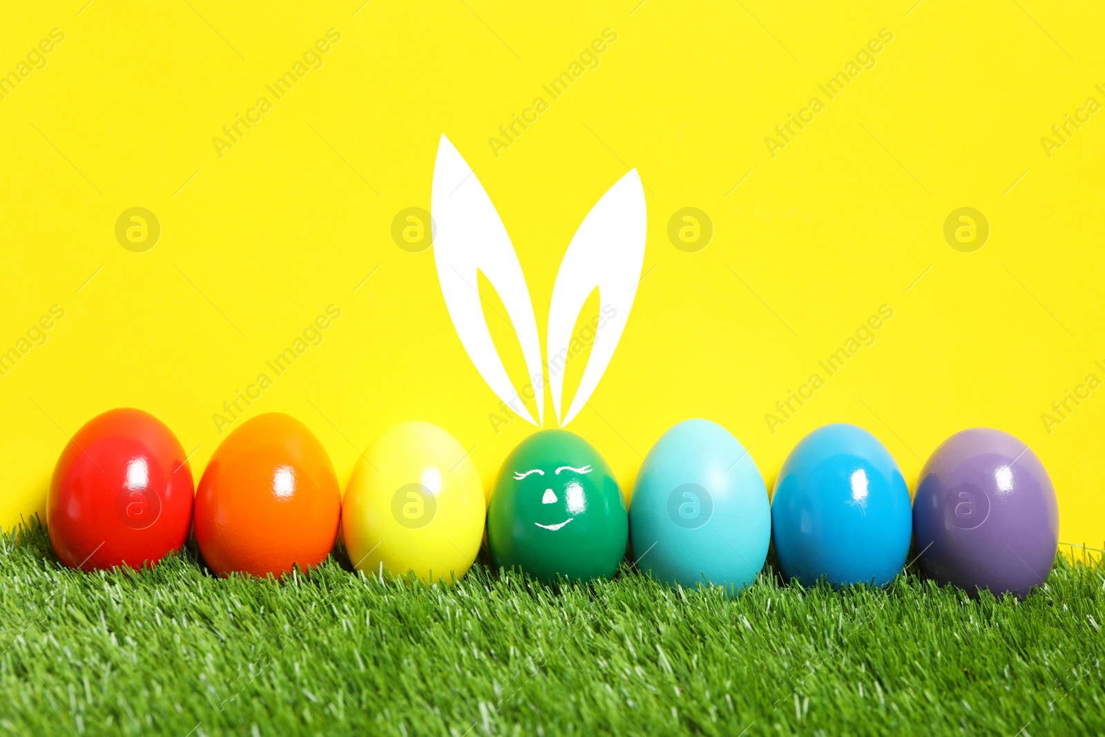 Image of One egg with drawn face and ears as Easter bunny among others on green grass against yellow background