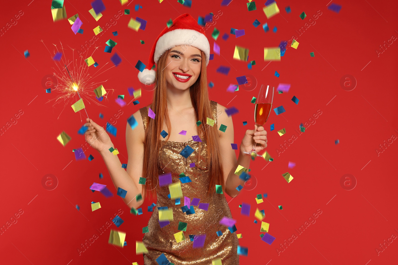 Image of Happy woman in Santa hat with sparkler and glass of sparkling wine under falling confetti on red background