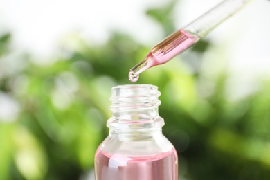 Dropper with essential oil over bottle against blurred background, closeup