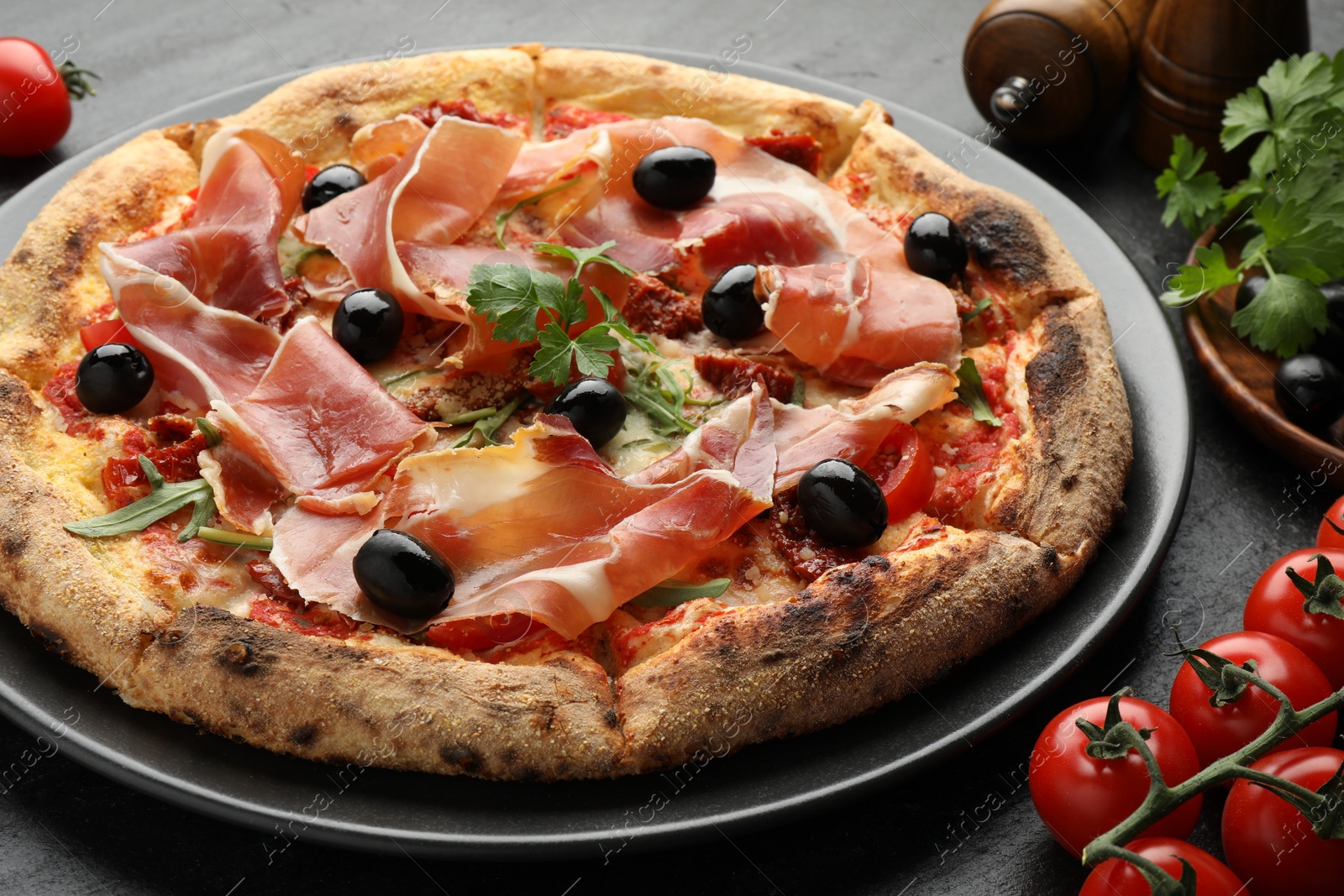 Photo of Tasty pizza with cured ham, olives, tomatoes and parsley on black table