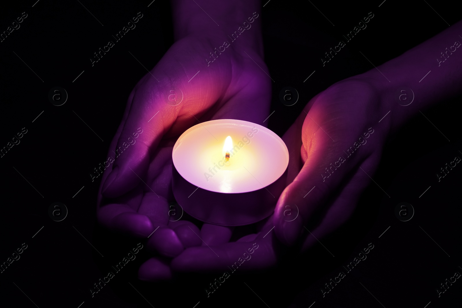 Image of Woman holding burning violet candle in hands on black background, closeup. Funeral attributes