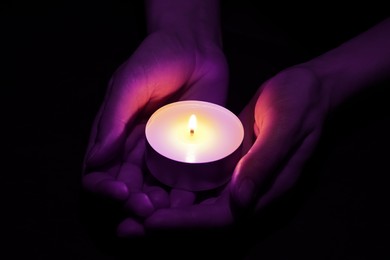 Woman holding burning violet candle in hands on black background, closeup. Funeral attributes