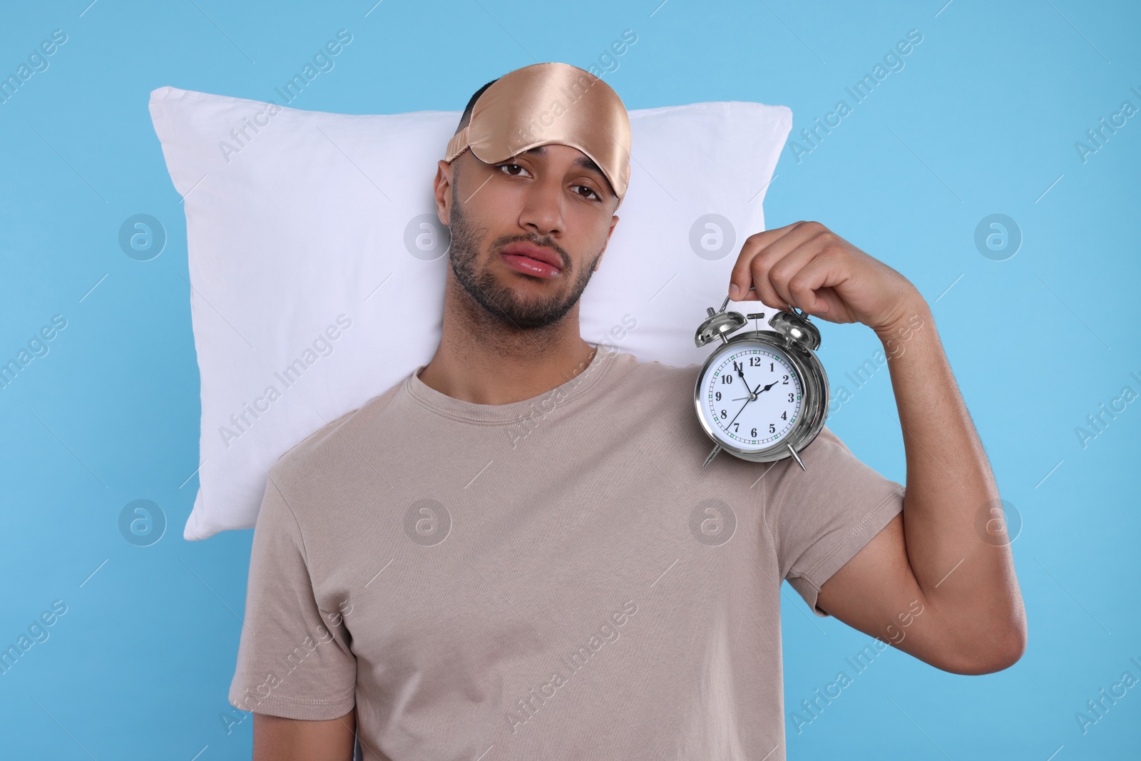 Photo of Tired man with pillow, sleep mask and alarm clock on light blue background. Insomnia problem