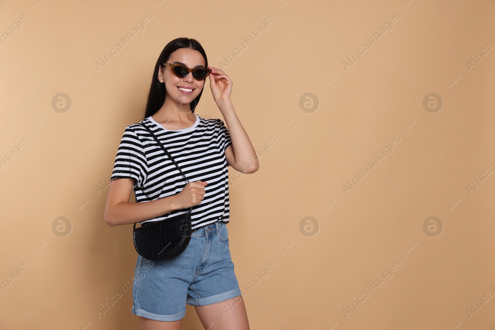 Photo of Beautiful young woman with stylish bag in sunglasses on beige background, space for text