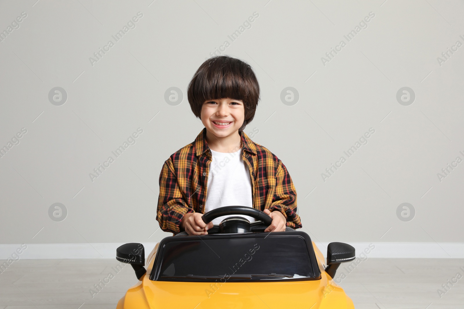 Photo of Cute little boy driving children's electric toy car near grey wall indoors