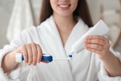 Photo of Woman squeezing toothpaste from tube onto electric toothbrush in bathroom, closeup