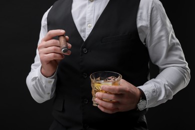 Man with glass of whiskey smoking cigar against black background, closeup