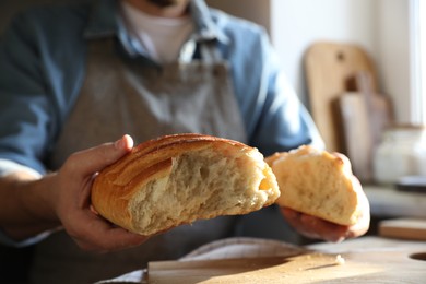 Photo of Man breaking loaf of fresh bread at wooden table indoors, closeup