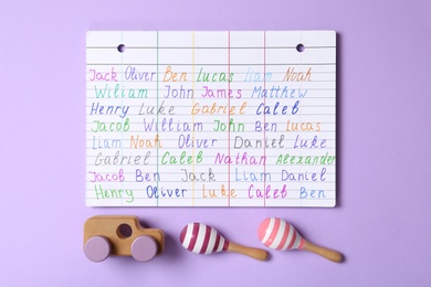 Photo of Paper sheet with written different baby names and wooden toys on purple background, flat lay
